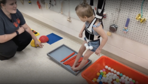 A young child in a HOPSA dress uses their feet to touch items on a sensory board below as a teacher looks on.